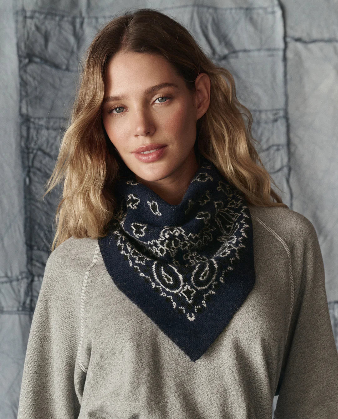 A woman with wavy, light brown hair wears a grey top and The Bandana Kerchief by The Great Inc.—a dark blue alpaca accessory with a white paisley design—around her neck as she stands in front of a textured grey fabric background.
