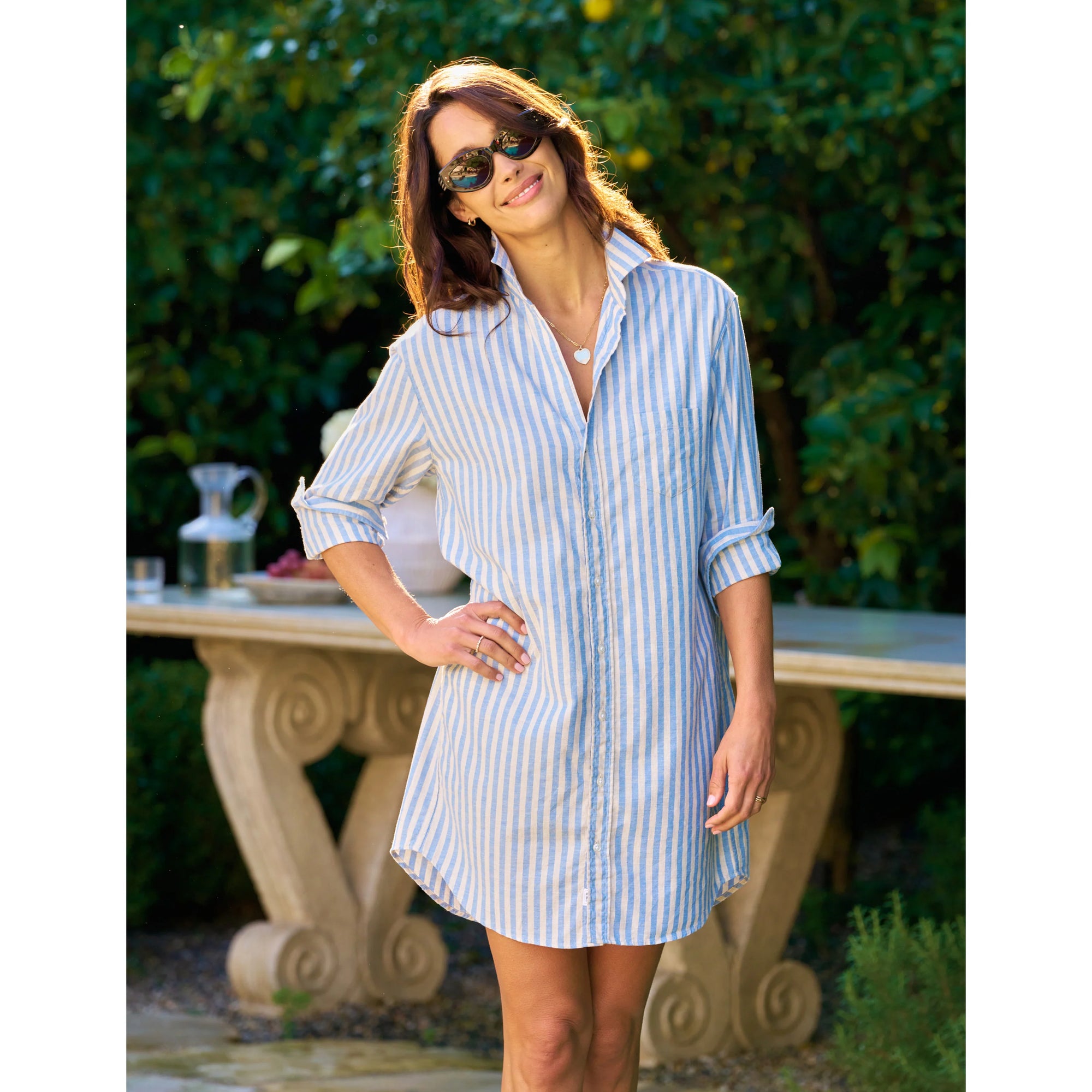 A woman wearing sunglasses and the Frank &amp; Eileen MARY Shirtdress S25 stands outdoors, smiling with one hand on her hip. Green foliage and a stone table in the background highlight this fresh look.