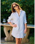 A woman wearing sunglasses and the Frank & Eileen MARY Shirtdress S25 stands outdoors, smiling with one hand on her hip. Green foliage and a stone table in the background highlight this fresh look.