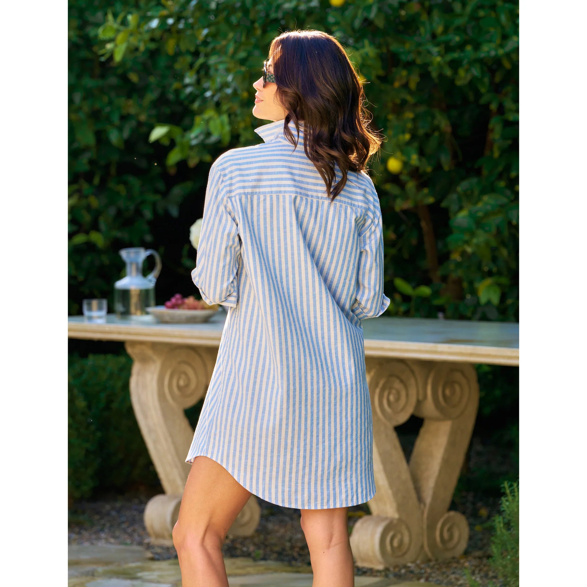 A woman with wavy brown hair stands outdoors, facing away, wearing the Frank &amp; Eileen MARY Shirtdress S25 in blue and white stripes. Behind her, a stone table holds a glass pitcher and fruit amid lush greenery.
