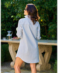 A woman with wavy brown hair stands outdoors, facing away, wearing the Frank & Eileen MARY Shirtdress S25 in blue and white stripes. Behind her, a stone table holds a glass pitcher and fruit amid lush greenery.