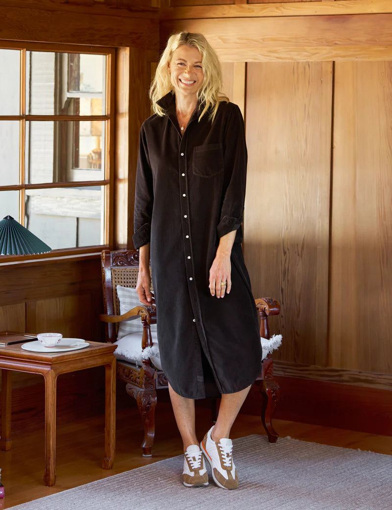 A woman with long blonde hair smiles while standing indoors in a warmly lit room. She is wearing the Rory Shirtdress S24 by Frank & Eileen and white sneakers. Behind her are wooden walls, a window, a chair, and a small table with a teacup.