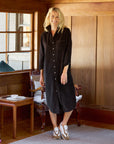 A woman with long blonde hair smiles while standing indoors in a warmly lit room. She is wearing the Rory Shirtdress S24 by Frank & Eileen and white sneakers. Behind her are wooden walls, a window, a chair, and a small table with a teacup.