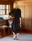 A woman with wavy blonde hair stands in a cozy, wooden-paneled room. She is wearing the Rory Shirtdress S24 by Frank & Eileen, a loose-fitting black maxi dress with rolled-up sleeves, paired with white sneakers. In the background, there is a chair, a side table holding a hardcover book and a cup, along with a lamp and window.