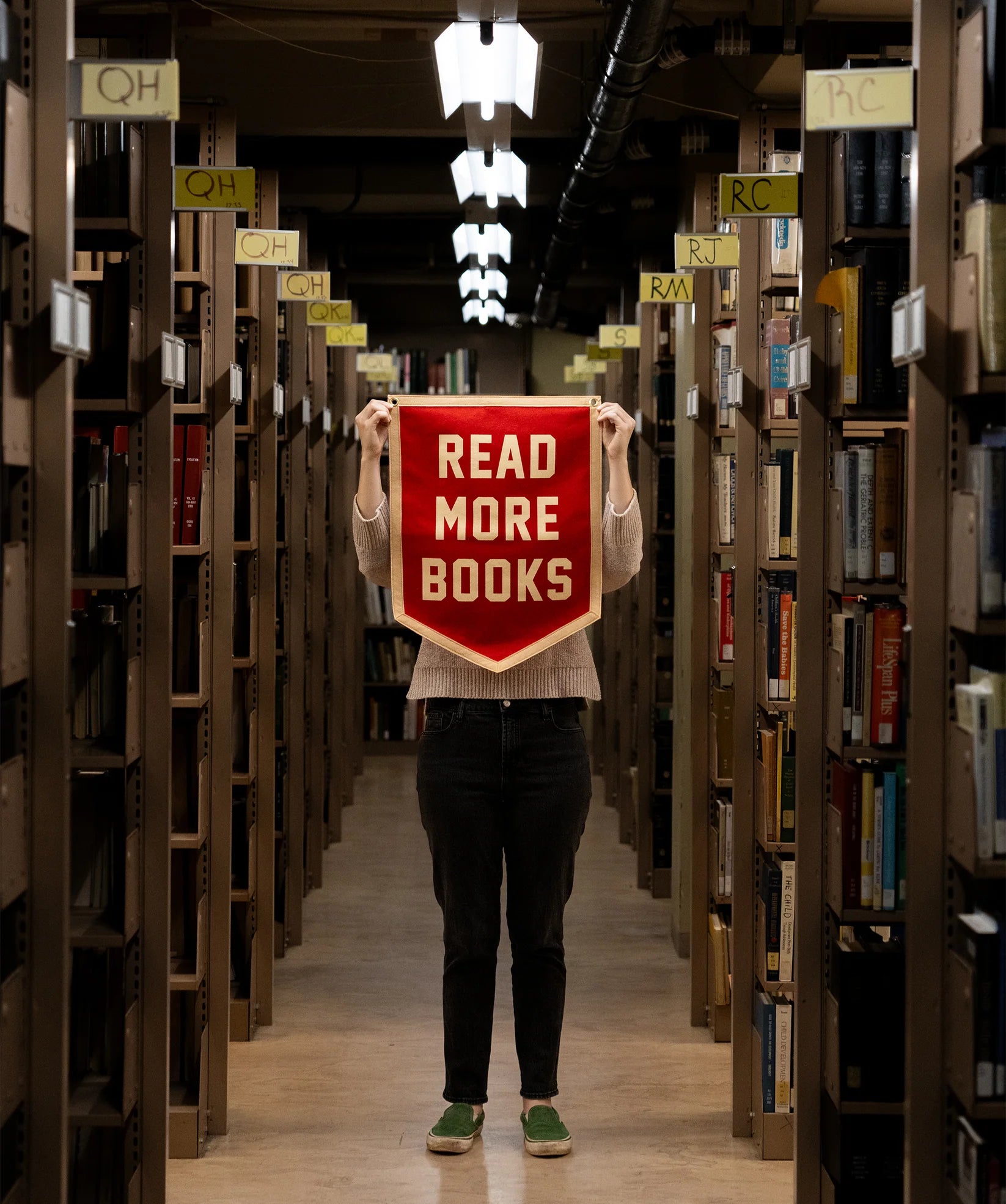 A person stands between library shelves, holding an Oxford Pennant "Read More Books" 18x24 pennant, with only their arms and legs visible. Call number signs hang above the shelves, celebrating a love of books.