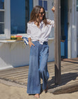 A woman wears Frank & Eileen's Madeline Patch Pocket Pant, a pair of indigo denim wide leg pants, with a white shirt as she stands barefoot on a wooden beach house deck, smiling and holding a post with food and a towel in the background.