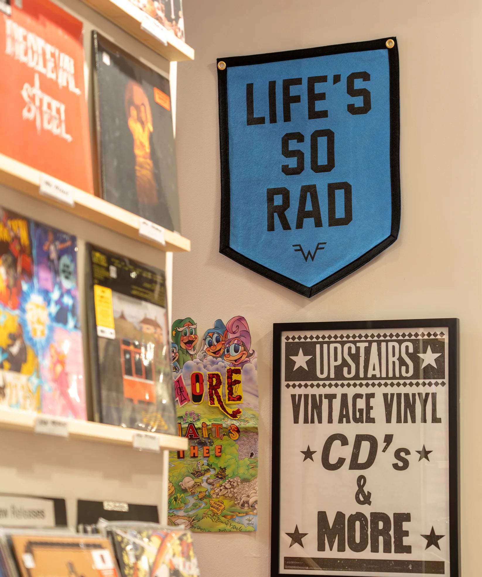 A record store wall features Oxford Pennant’s bright blue “Life’s So Rad” flag, shelves of vinyl, a vibrant cartoon poster, and a black-and-white sign for vintage vinyl and CDs upstairs—ideal dorm room decor inspo.