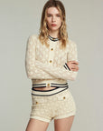 A woman with long blonde hair poses against a plain light background, arms crossed, wearing the FRAME Rookie Cardi—a lightweight knit cardigan styled with matching high-waisted shorts featuring gold buttons and black trim.