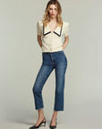 A woman stands against a plain background wearing a cream blouse with a bold black-trimmed collar, FRAME's The Ruler Crop slim-straight jeans in medium-blue with frayed hems, and black heels. Her hands are in her pockets as she gazes forward.
