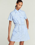 A woman stands against a plain background wearing FRAME’s Striped Safari Dress—a light blue, short-sleeve cotton poplin dress with white vertical pinstripes, front pockets, and a matching belted waist.