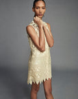 A woman wears the FRAME Deco Lace Dress, a sleeveless, light-toned piece inspired by 1920s shift silhouettes and intricate lace, standing against a plain gray backdrop while gazing at the camera and touching her neck.