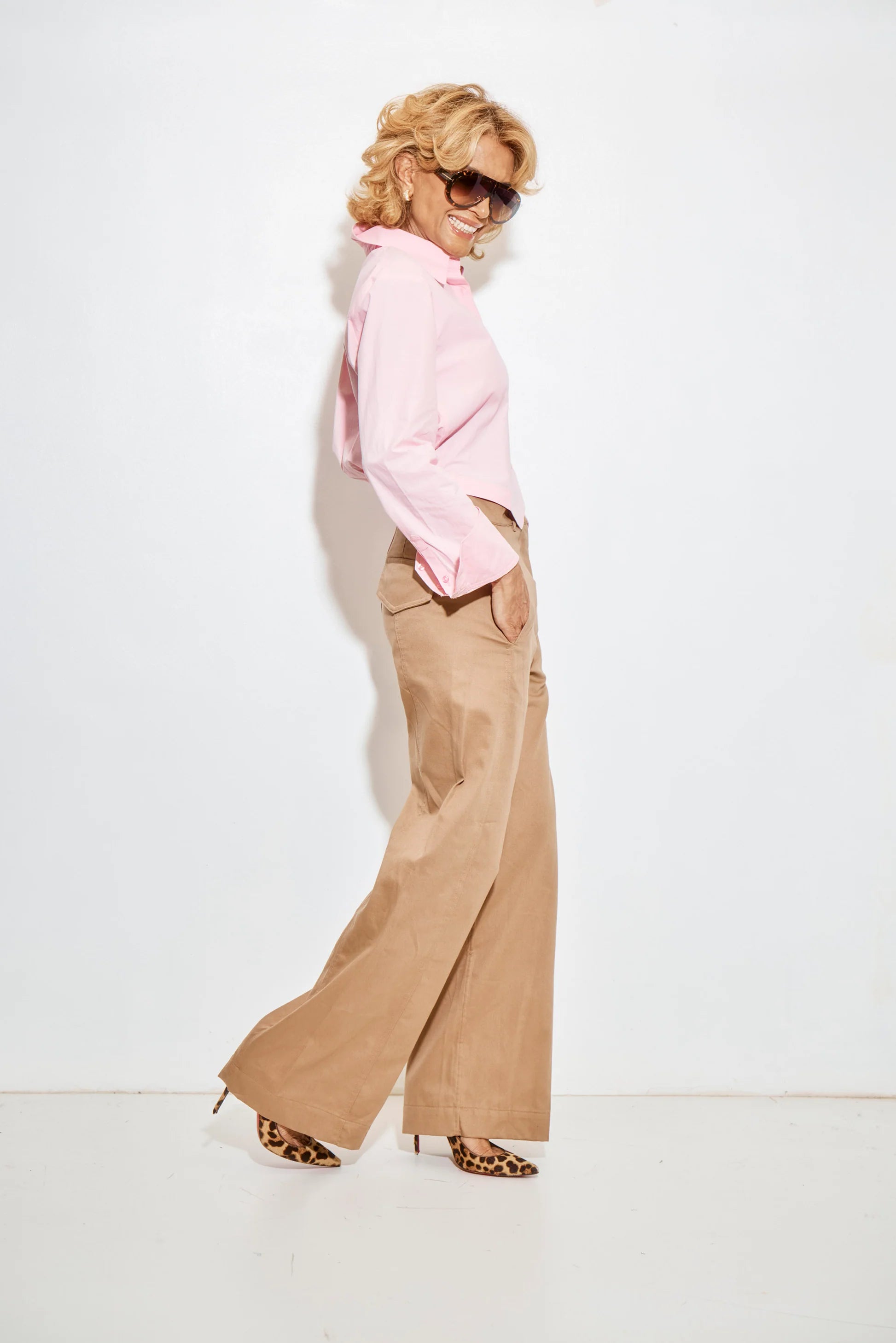 A stylish woman with blond hair models the Colette Parachute button-down shirt in light pink by A Shirt Thing, paired with tan wide-leg pants and leopard print heels. She smiles, posing sideways with hands in her pockets against a white background.