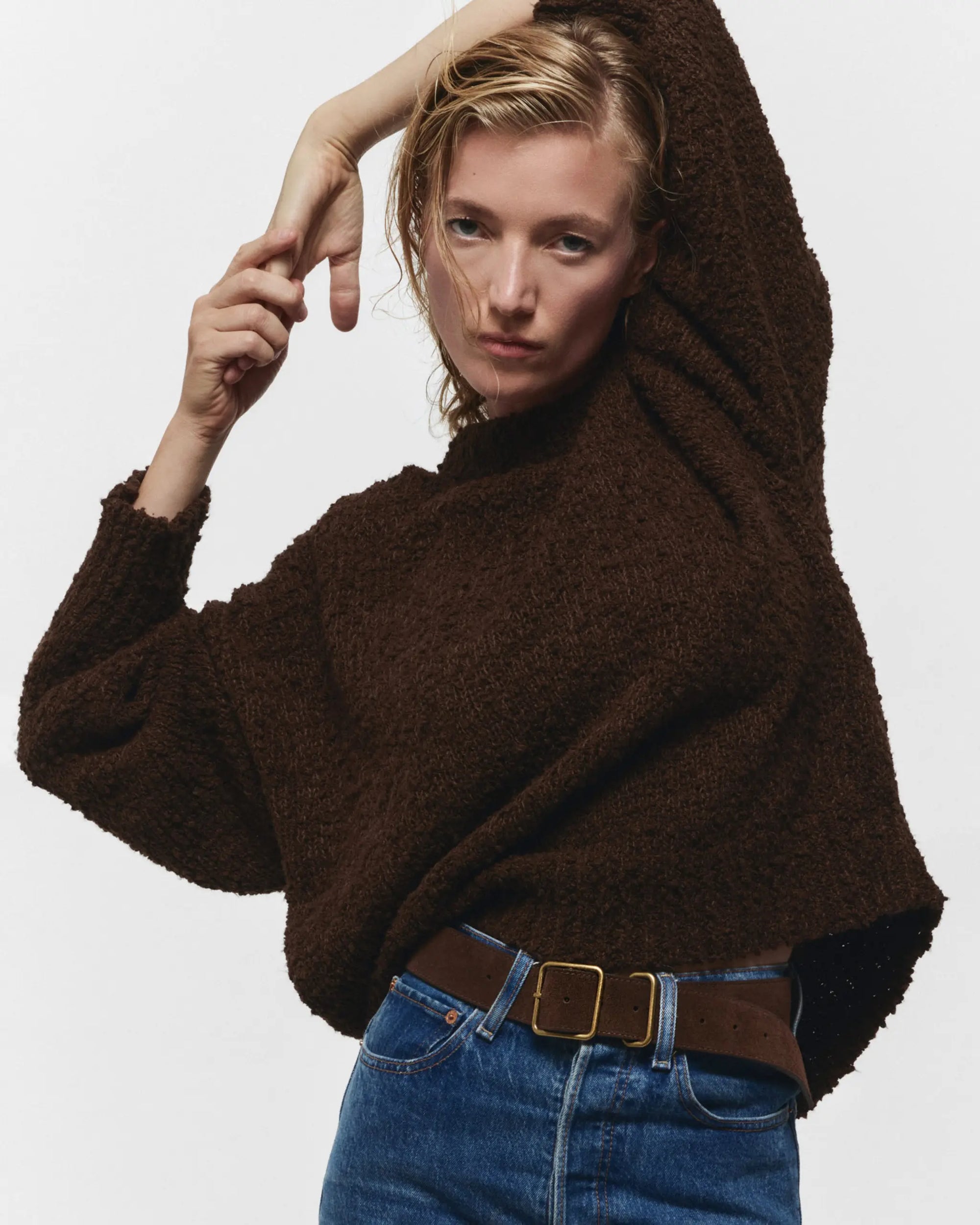 A person models the Janessa Leone Bowe Belt, styled with a textured dark brown sweater and blue jeans, striking a confident pose against a plain white background.