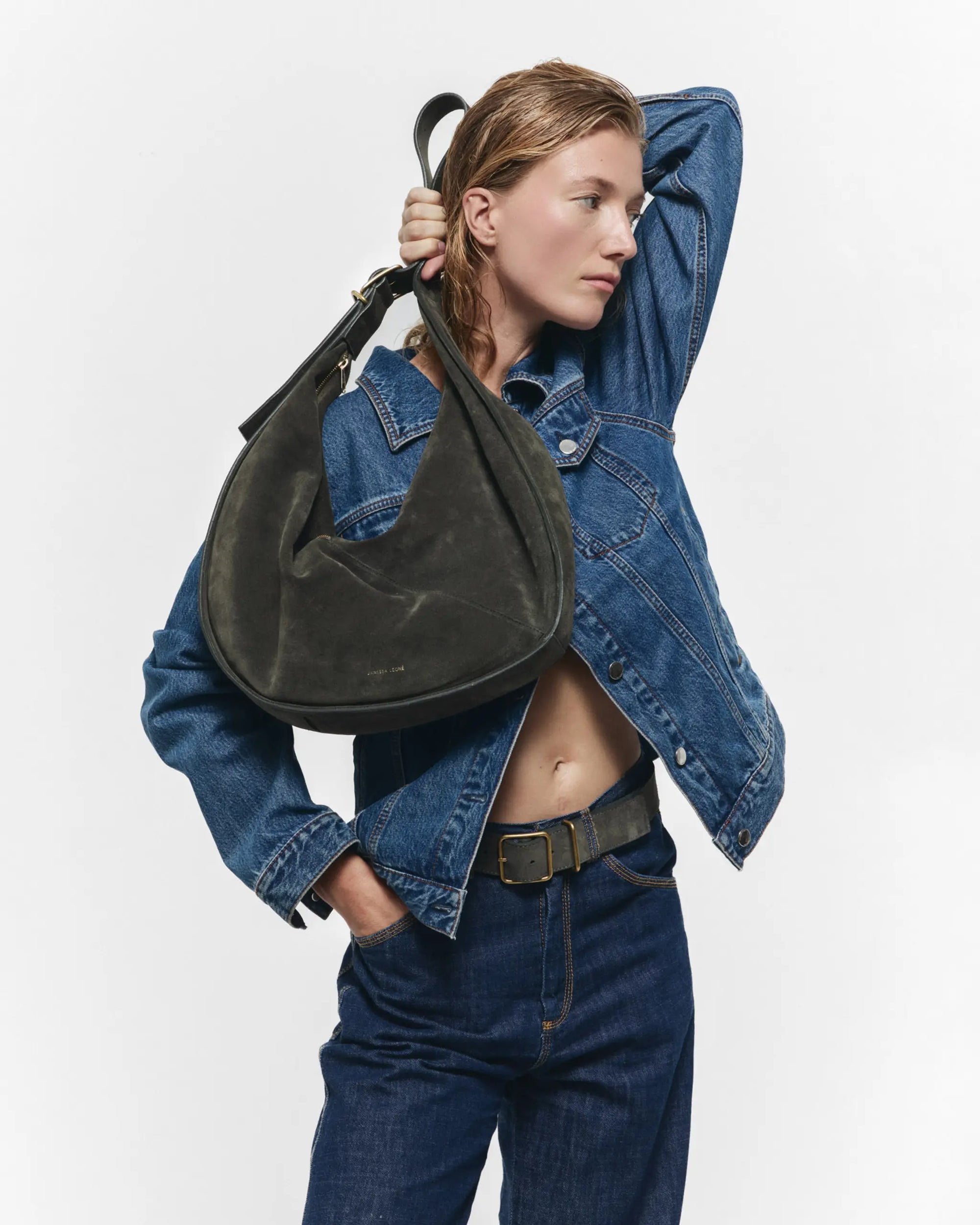 A person in a blue denim jacket and jeans poses with one hand behind their head, showcasing the Janessa Leone Bowe Belt over their shoulder against a plain white background.