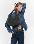 A person in a blue denim jacket and jeans poses with one hand behind their head, showcasing the Janessa Leone Bowe Belt over their shoulder against a plain white background.