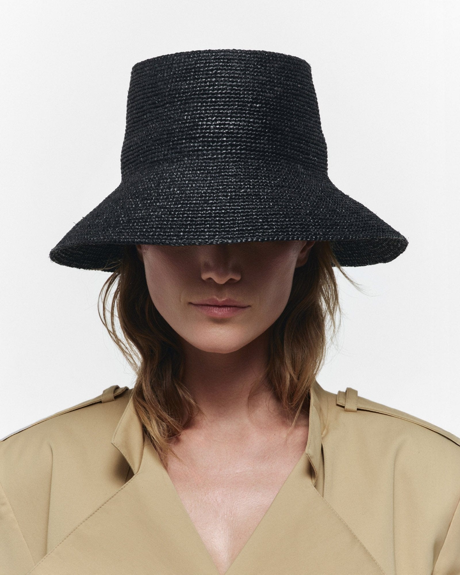 A woman wears the Janessa Leone FELIX BUCKET wide-brimmed black straw hat, paired with a beige trench coat against a plain white background.