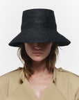 A woman wears the Janessa Leone FELIX BUCKET wide-brimmed black straw hat, paired with a beige trench coat against a plain white background.