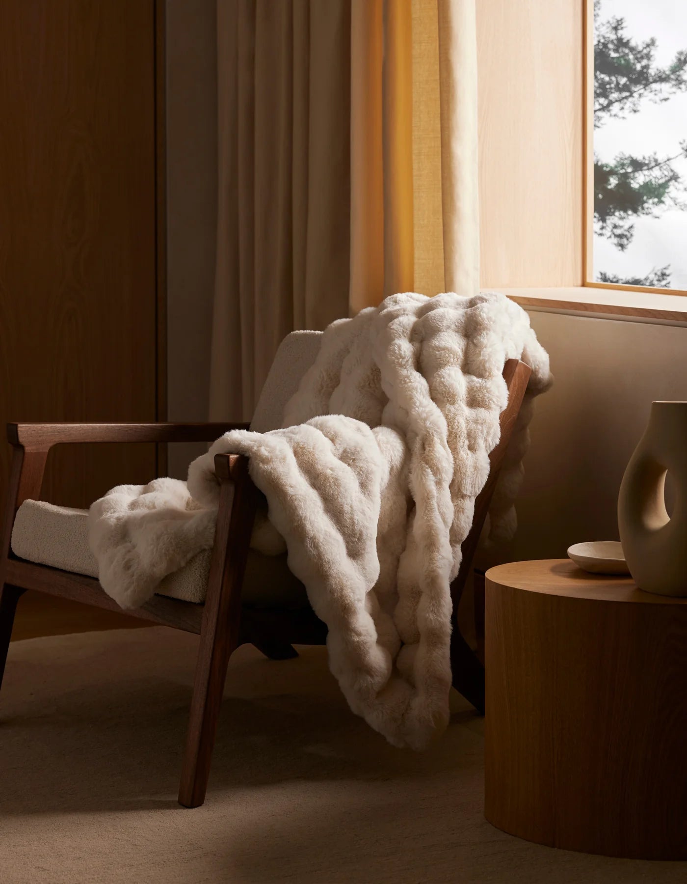 The Cozy Earth Bubble Cuddle Blanket, in creamy ultra-soft fabric, is draped over a wooden armchair by a window with beige curtains. Nearby, a round wooden side table with a decorative vase sits bathed in warm natural light.