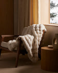 The Cozy Earth Bubble Cuddle Blanket, in creamy ultra-soft fabric, is draped over a wooden armchair by a window with beige curtains. Nearby, a round wooden side table with a decorative vase sits bathed in warm natural light.