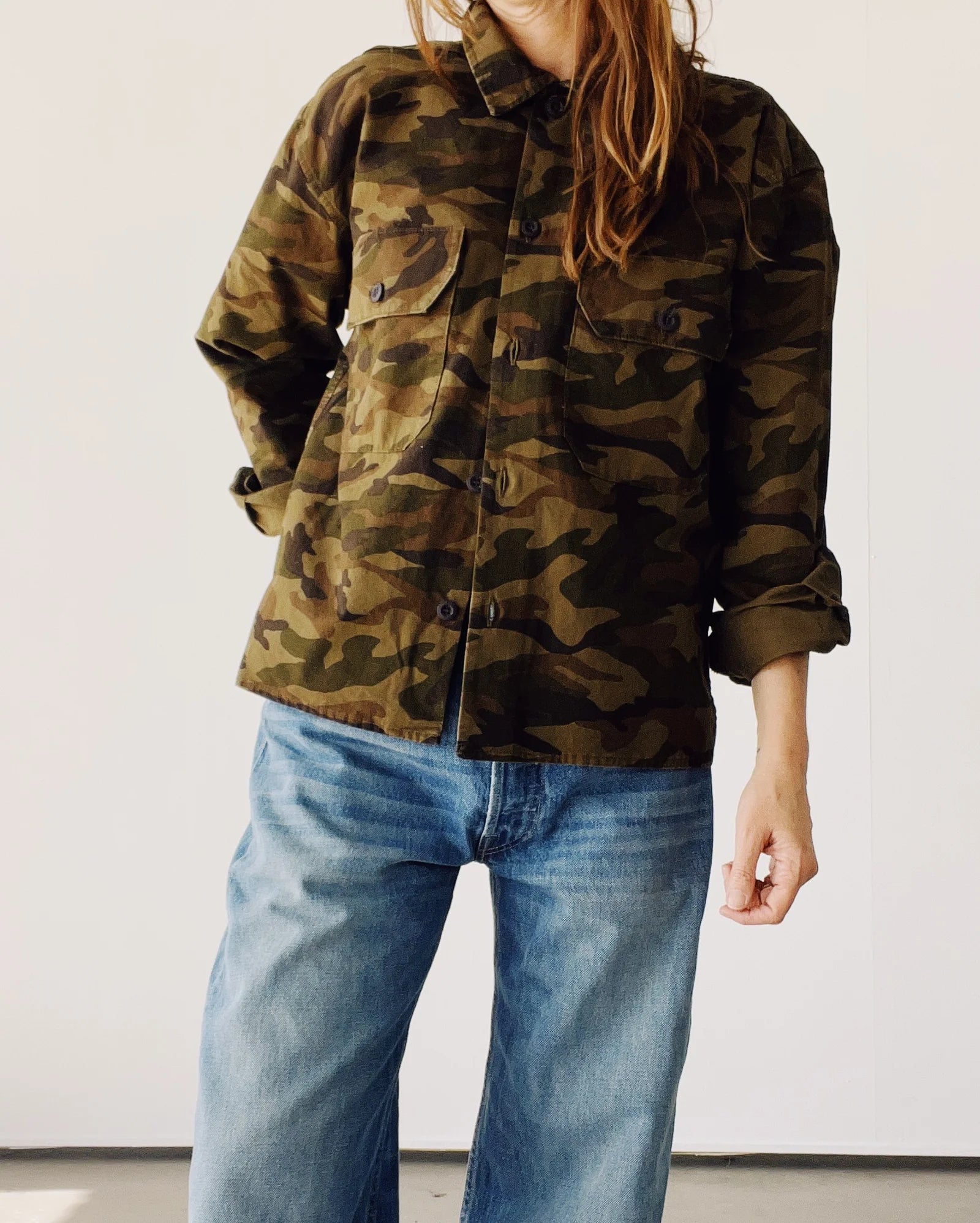 Model wearing The Work Shirt by Hey Gang in a boxy fit camouflage design with rolled-up sleeves, paired with blue jeans. Photographed against a plain light background; head is partially out of frame.