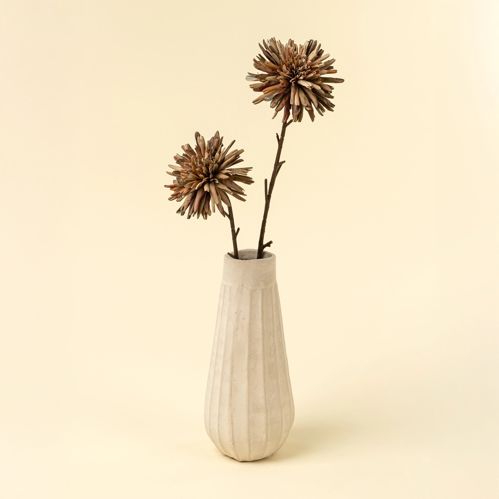 A white, textured ceramic vase holds two Indaba Dahlia Stem Russet faux flowers with long, slender stalks, serving as minimal accent decor against a plain, light beige background.