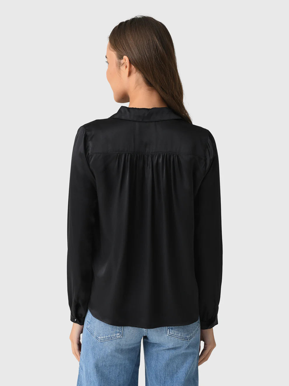 A woman with long brown hair is seen from behind, modeling the Celine Silk black shirt with ruffles by A Shirt Thing and light blue jeans, set against a plain light gray background.