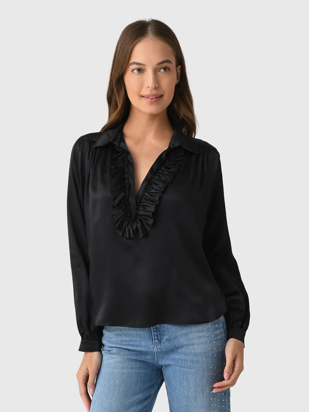 A woman with long brown hair wears the Celine Silk shirt by A Shirt Thing, a black long-sleeve silk top with cascading ruffles along the V-neckline, paired with blue jeans. She stands against a light gray background, smiling softly.
