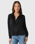 A woman with long brown hair wears the Celine Silk shirt by A Shirt Thing, a black long-sleeve silk top with cascading ruffles along the V-neckline, paired with blue jeans. She stands against a light gray background, smiling softly.