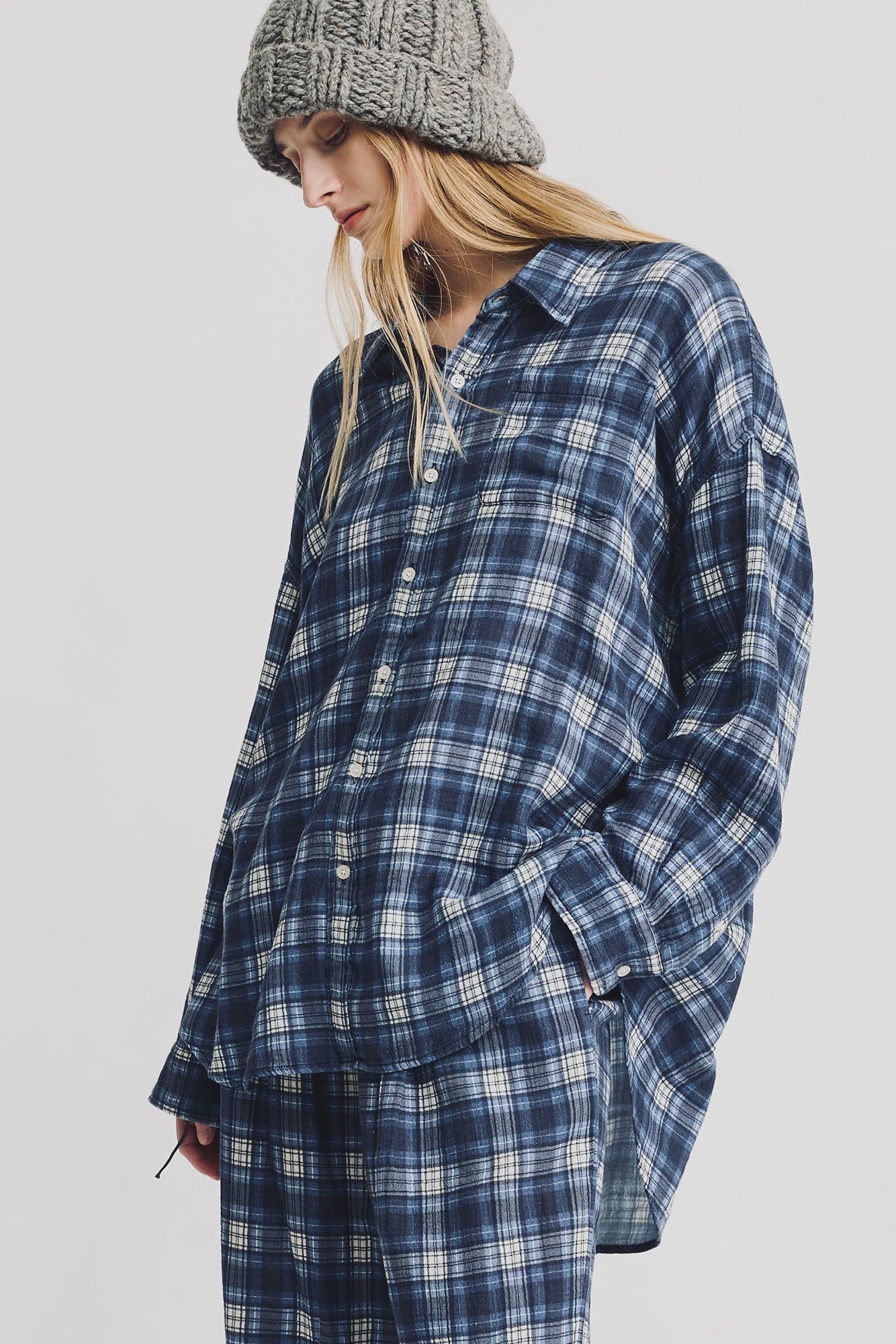 A woman with long blonde hair wears the Denimist BUTTON FRONT SHIRT in blue/ecru plaid, styled with matching pants and a chunky gray knit beanie. She stands against a white background with one hand in her pocket.