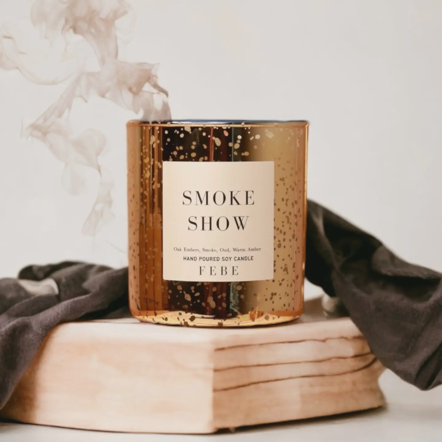 A lit golden FEBE Candle by Faire exudes a smokey fragrance with hints of oud, sitting on a wooden block amid wisps of smoke. It's nestled in dark cloth, crafting a cozy ambiance.