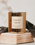 A lit golden FEBE Candle by Faire exudes a smokey fragrance with hints of oud, sitting on a wooden block amid wisps of smoke. It's nestled in dark cloth, crafting a cozy ambiance.