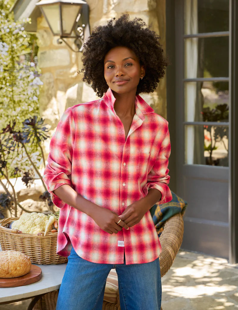A woman stands outside by a stone wall and glass door, smiling gently in the Frank & Eileen Eileen Button Up Shirt F25, a red and white plaid style, paired with blue jeans. A basket of bread and vegetables sits on a nearby table.