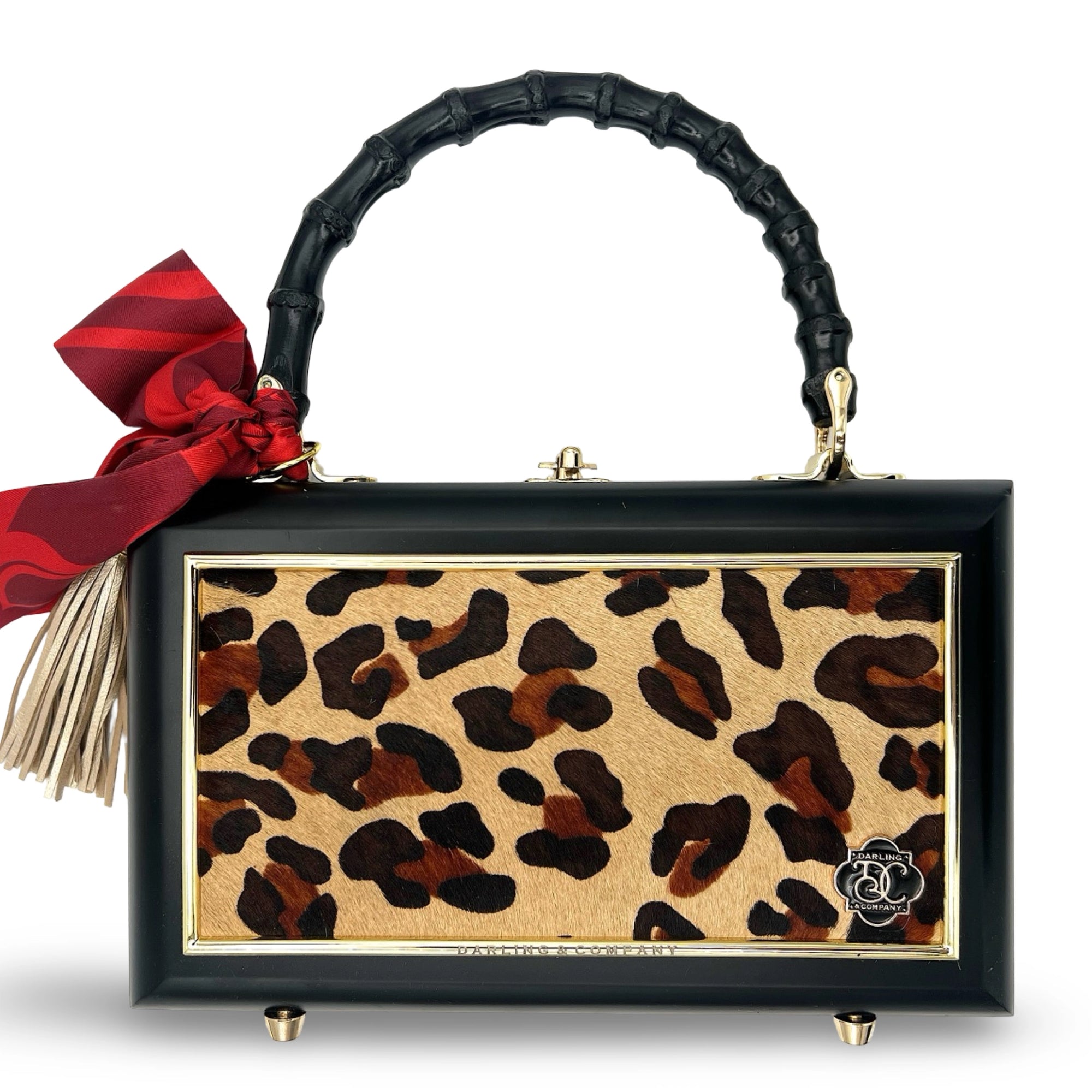 The Faire Leopard Supreme Bag is a rectangular leopard print cowhide purse with a black frame, gold accents, black bamboo handle, red bow, beige tassel, and four metal feet for added sophistication.