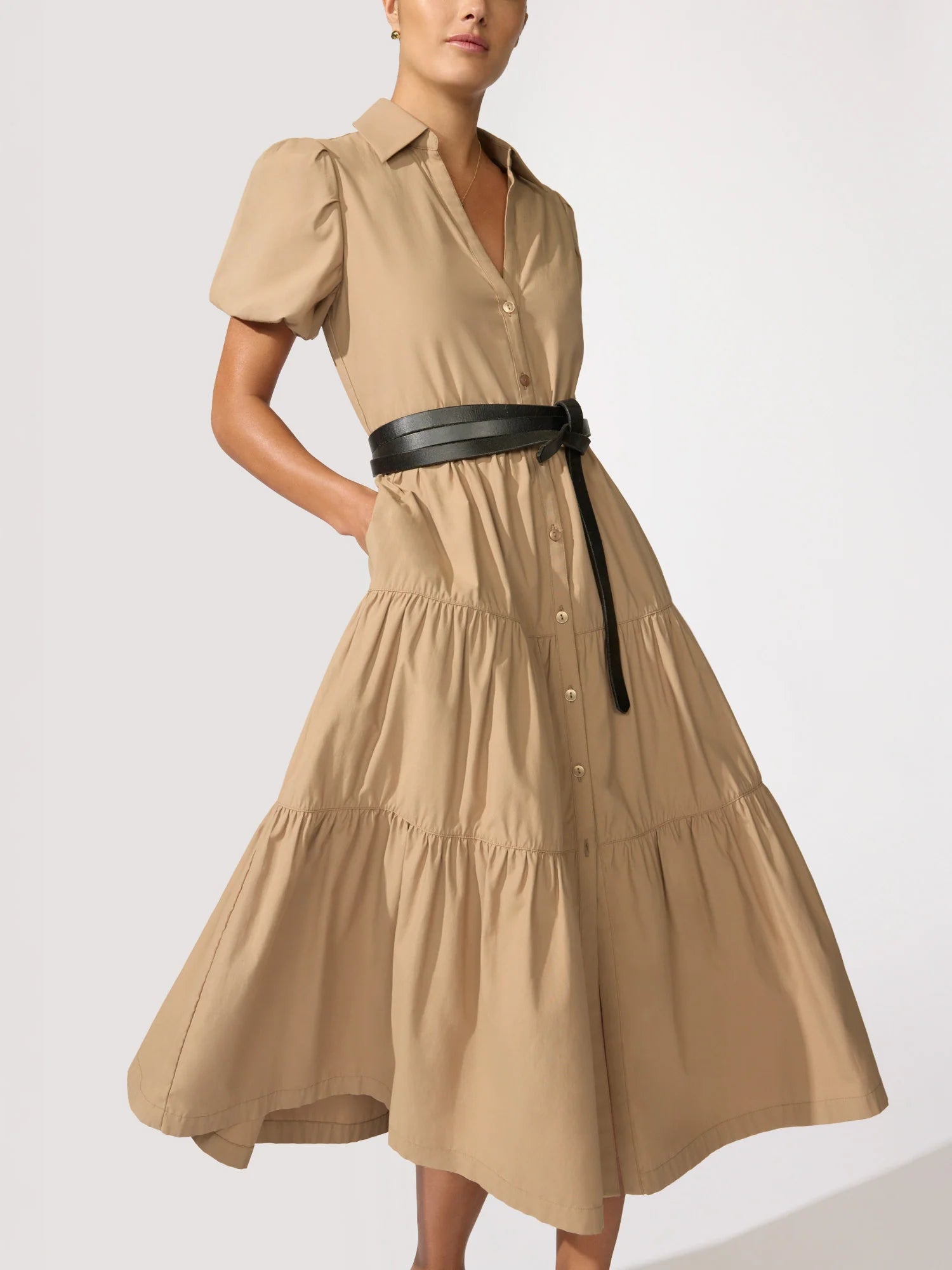 A woman models the HAVANA DRESS by BROCHU WALKER, a tan, short-sleeve, button-down dress with a tiered skirt and pockets. She stands against a light background, her waist cinched with a black belt.