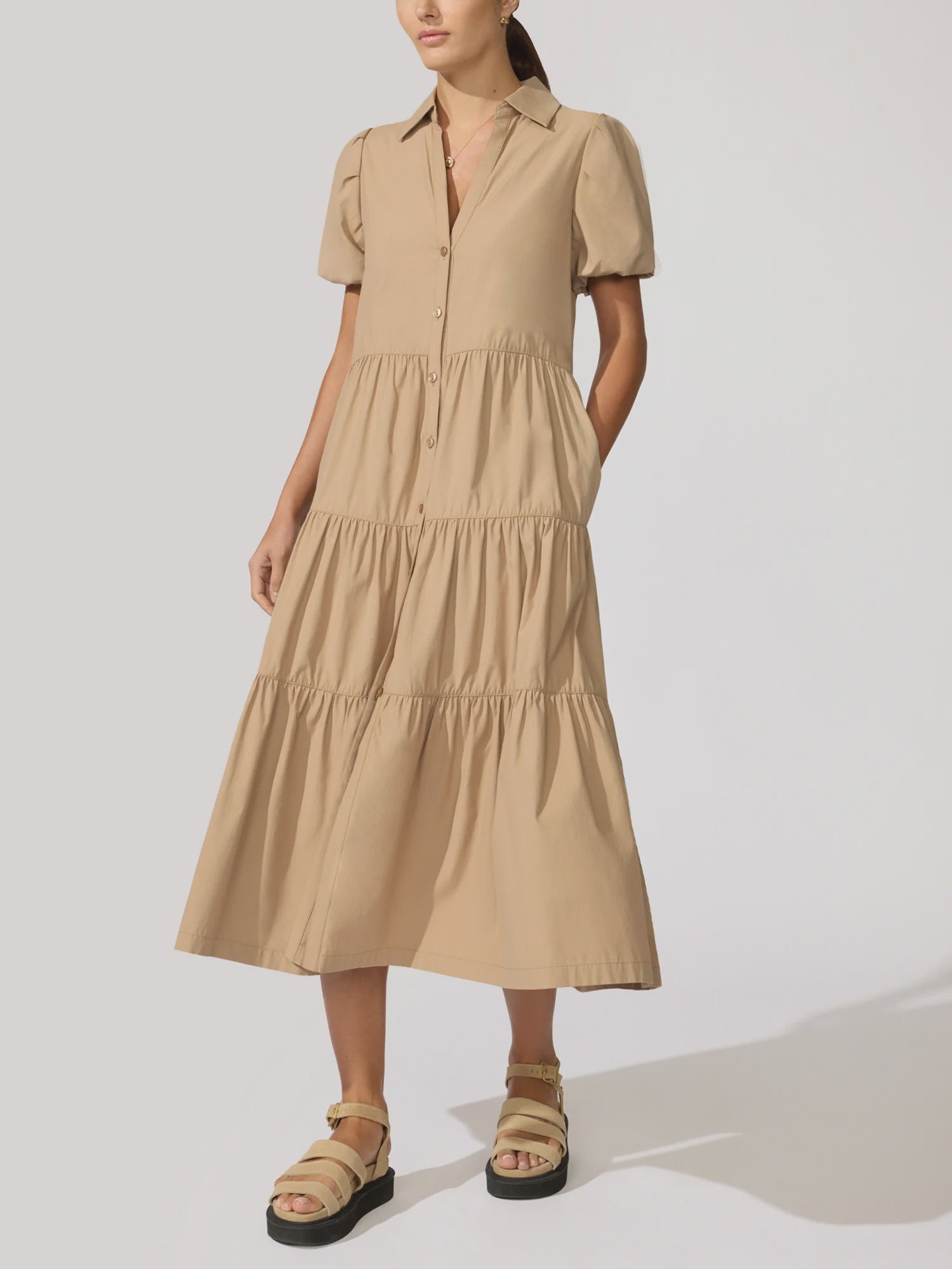 A woman wears the BROCHU WALKER HAVANA DRESS, featuring a tan hue, tiered skirt, button front, and collar. She pairs it with beige sandals and stands against a plain light background, one hand in her pocket.