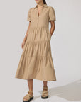 A woman wears the BROCHU WALKER HAVANA DRESS, featuring a tan hue, tiered skirt, button front, and collar. She pairs it with beige sandals and stands against a plain light background, one hand in her pocket.