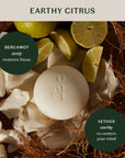 Faire's Grounding Stone Body Lotion, a round white luxury bar, is paired with bergamot (zesty, restores focus), lime slices, wax chunks, and vetiver (earthy, re-centers your mind).