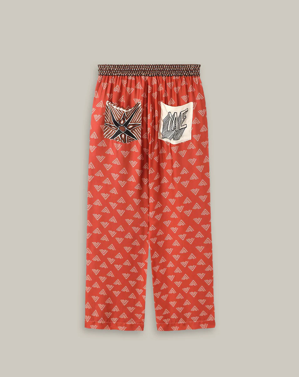 The Me369 Bailey Track Pant is loose-fitting and red-orange with a white geometric pattern, elastic waistband, and two large back pockets featuring abstract black-and-white graphic designs.