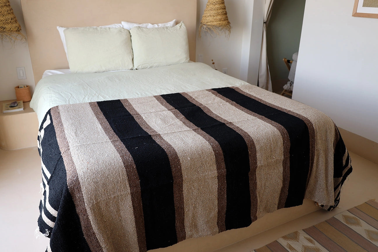 Faire Blackstone Lines Handwoven Blanket on a bed in a cozy room with light pillows and straw lamps, Bungalow style.
