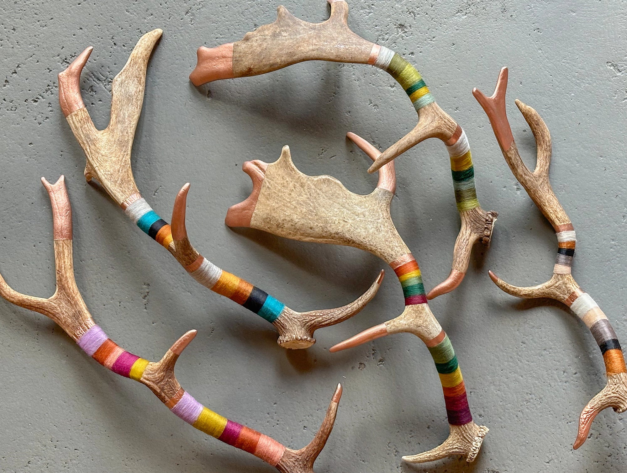 The Faire Wool & Copper Wrap Deer Antler Fallow features handcrafted antlers on a gray surface, each partly wrapped in colorful yarn, creating vibrant, unique wall decor.