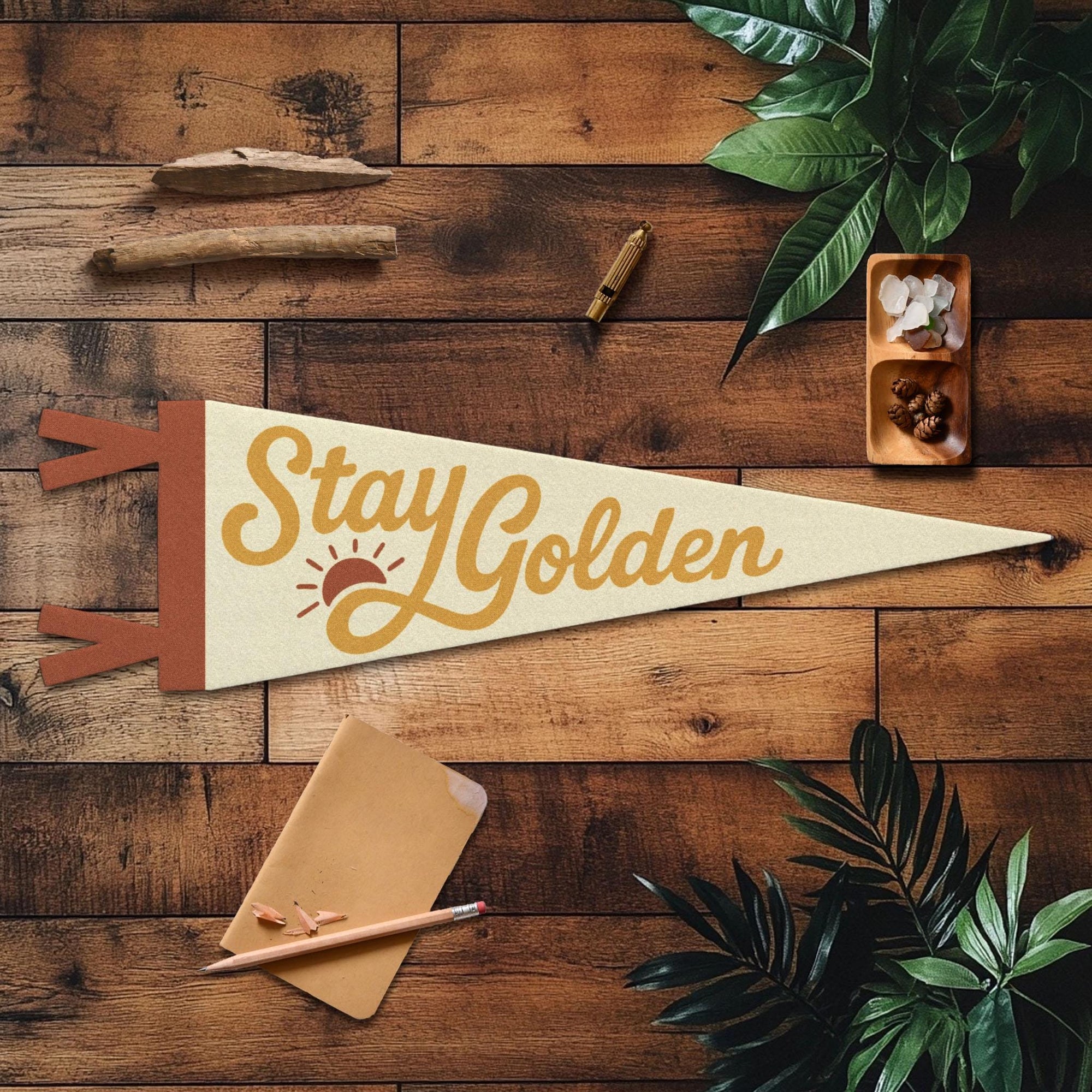 The "Stay Golden" Felt Pennant by Faire is a handmade wall decor featuring the phrase and a sun graphic, displayed on a wooden surface with green leaves, notebook, pencil, crystals, and a cigar.