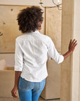 A woman with curly hair stands indoors, seen from behind, wearing the Frank & Eileen BARRY Tailored white button-up shirt with rolled sleeves and blue jeans, resting one hand on a beige column as she faces an abstract wall painting.