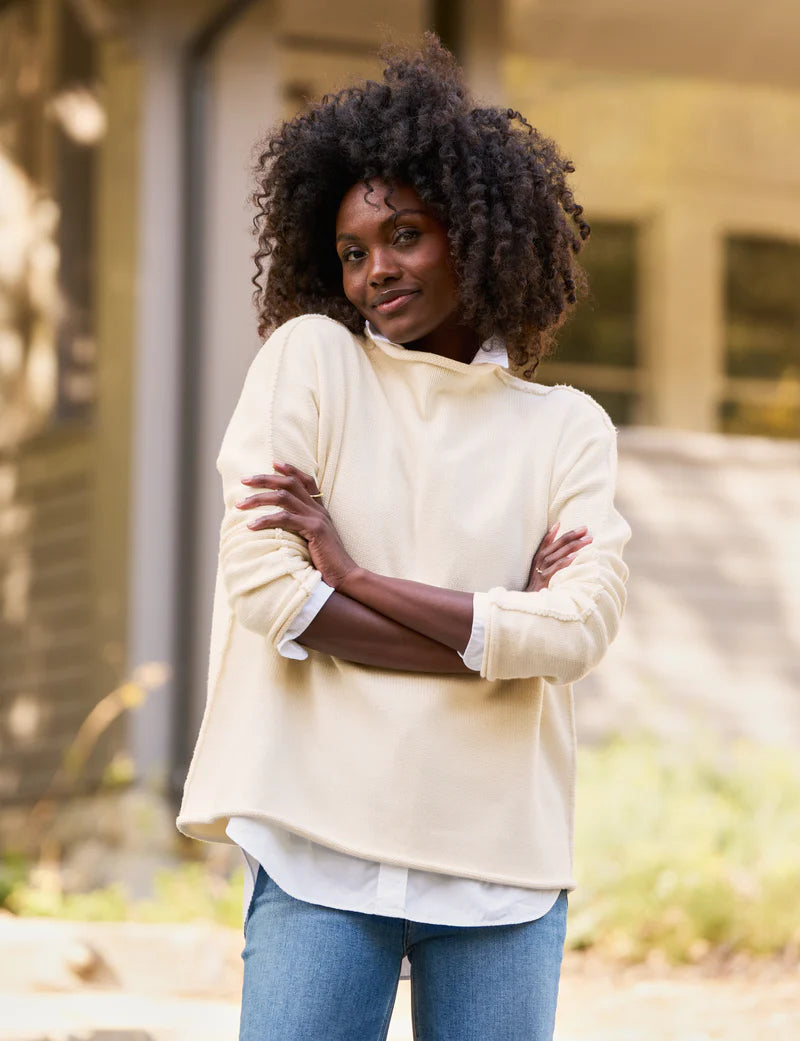 A woman with curly hair, wearing the luxurious Monterey Sweater from Frank &amp; Eileen and blue jeans, stands outdoors with her arms crossed, smiling softly. The background is blurred, featuring part of a building and greenery. Her sweater, knitted by artisans from Italian cotton, adds an elegant touch to the scene.