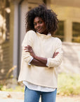 A woman with curly hair, wearing the luxurious Monterey Sweater from Frank & Eileen and blue jeans, stands outdoors with her arms crossed, smiling softly. The background is blurred, featuring part of a building and greenery. Her sweater, knitted by artisans from Italian cotton, adds an elegant touch to the scene.