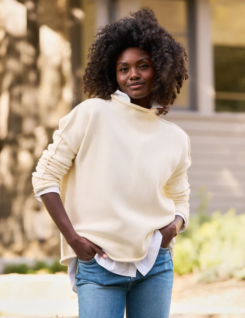 A person with curly hair stands outdoors, smiling slightly. They are wearing the luxurious Monterey Sweater by Frank &amp; Eileen, knitted by artisans, over a white shirt and blue jeans. The background is blurred, with hints of greenery and a building.