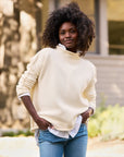 A person with curly hair stands outdoors, smiling slightly. They are wearing the luxurious Monterey Sweater by Frank & Eileen, knitted by artisans, over a white shirt and blue jeans. The background is blurred, with hints of greenery and a building.