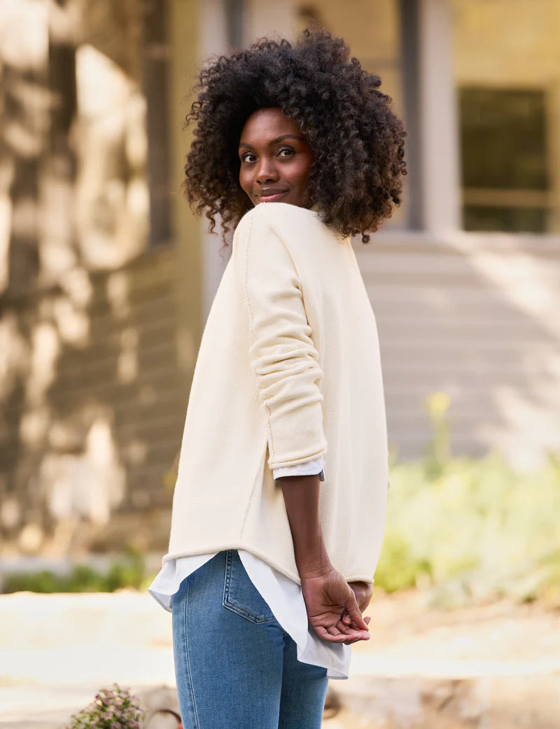 A person with curly hair, wearing a luxurious Monterey Sweater by Frank &amp; Eileen and blue jeans, stands outdoors, looking over their shoulder with a smile. The background is blurred, featuring trees and a building in the distance.