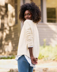 A person with curly hair, wearing a luxurious Monterey Sweater by Frank & Eileen and blue jeans, stands outdoors, looking over their shoulder with a smile. The background is blurred, featuring trees and a building in the distance.