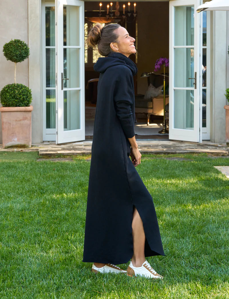 A woman in a maxi-length black Frank & Eileen James Hooded Cape Dress with a side slit and white sneakers stands on a grassy lawn, smiling and looking up, with open double doors and potted plants in the background.