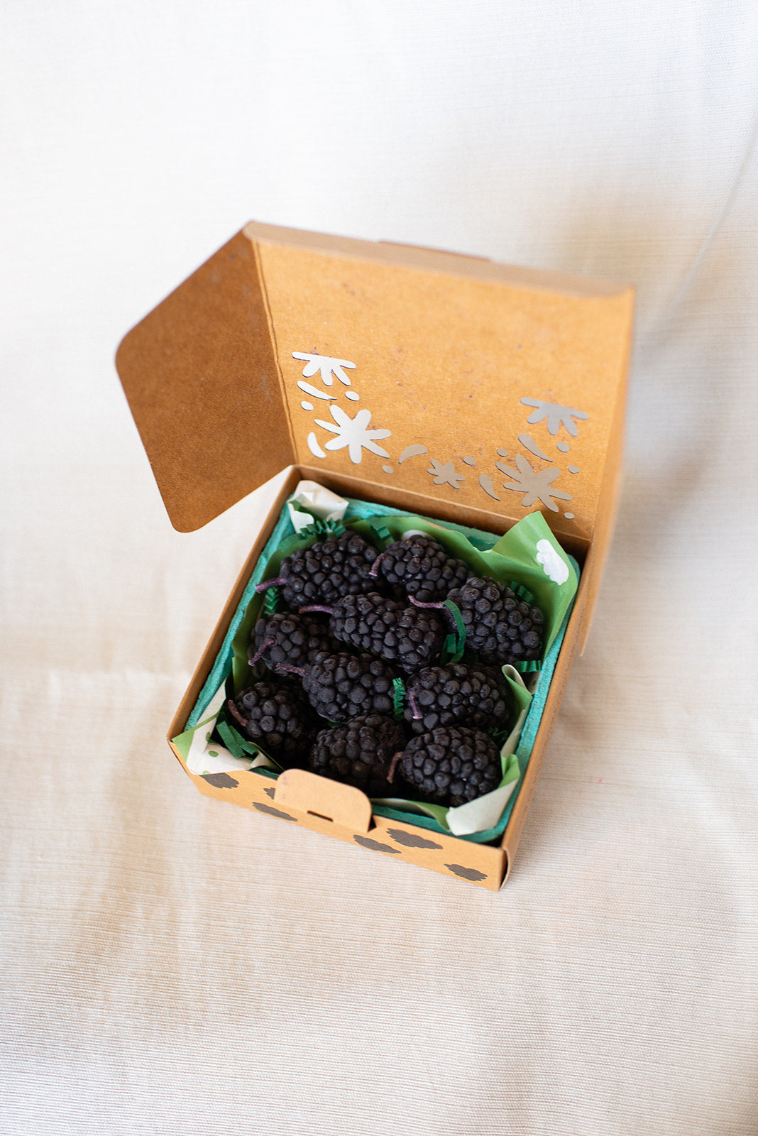 Faire’s Blackberry Beeswax Candles come in a small brown cardboard box with a floral cutout, nestled in green tissue paper. These blackberry-shaped candles add charm to any gift or celebration.